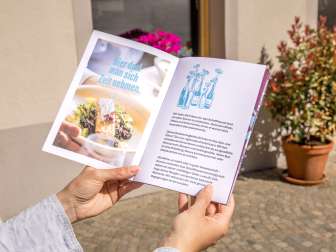 Eine Person hält das Genuss-Booklet in die Luft. Eine Broschüre mit Genusstipps und Gutscheinen zum Einlösen.
