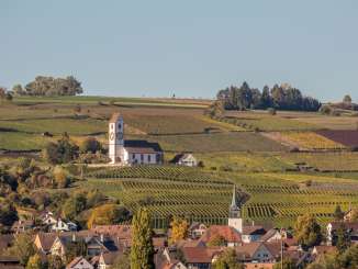 Im unteren Teil sind Häuser eines Dorfes. Weiter oben am Hang ist eine Kirche, umrundet von Weinreben, zu sehen.