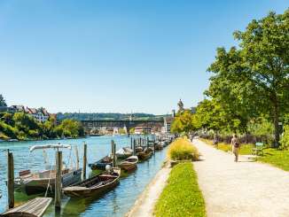 Flaniermeilie direkt am Rhein. Am Ufer stehen diverse Holzboote im Wasser und im Hintergrund sieht man die Festung Munot.
