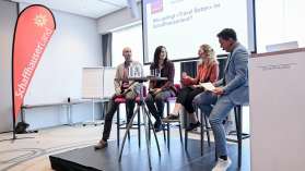 Vier Personen sitzen auf einer kleinen Bühne. Sie halten eine Podiumsdiskussion.