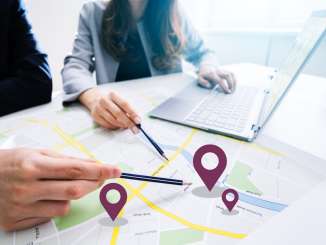 Zwei Personen sitzen an einem Tisch vor einem Laptop und einer ausgedruckten Karte. auf der Karte sind drei Standorte eingezeichnet.