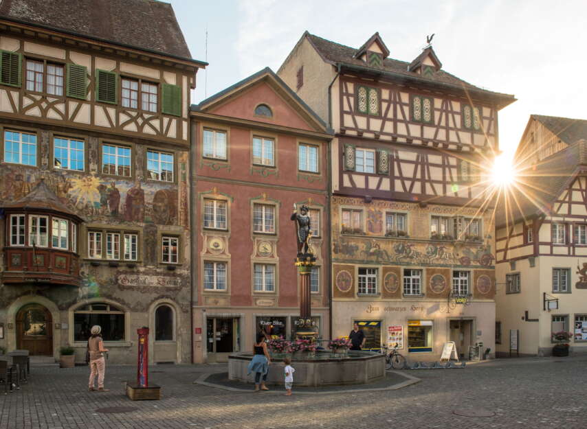 Fassadenmalereien in der Altstadt Stein am Rhein. Die Sonne scheint durch die Häuser. Vor den Häusern steht ein Brunnen.