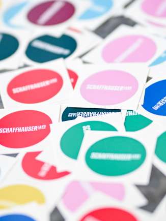 Es liegen viele bunte und runde Sticker auf einem Tisch. Auf den farbigen Hintergründen ist das Logo des Schaffhauserlandes.