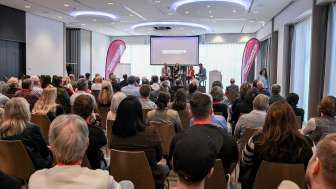 In einem Saal sitzen über 100 Personen. Auf einer kleine Bühne vor referieren vier Personen.
