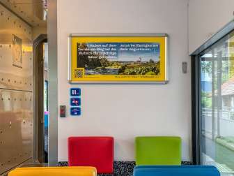 im Zug hängt ein Poster. Dieses hat zwei Bilder umrahmt von einem gelben Rahmen. Die Bilder zeigen einmal ein Bach/Fluss und einmal Reben mit einer Kirche.