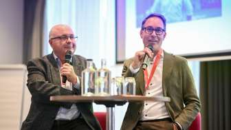 Zwei Männer sitzen auf einer kleinen Bühne. Sie halten ein Mikrofon in der Hand.