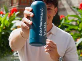 Ein Mann steht vor einem Brunnen in Stein am Rhein. In der Hand ausgestreckt hält er eine petrol-farbige Trinkflasche auf der "Schafuuserland, ich liebs." steht.