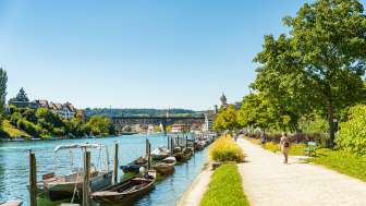 Flaniermeilie direkt am Rhein. Am Ufer stehen diverse Holzboote im Wasser und im Hintergrund sieht man die Festung Munot.