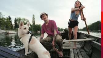 Eine Person und ein Hund sitzen in einem Holzboot. Eine Person steht und rudert und steuert den Weidling auf dem Rhein.