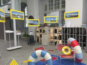 Auf einer leeren Fläche im Visitor Centre Schaffhausen ist eine kleine Ausstellung. Auf einem blauen Teppich stehen zwei rote und ein gelber Stuhl. Es liegen mehrere Schwimmreifen und Wasserbälle herum. Von einem Rind an der Decke hängen mehrere Schilder