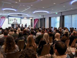 Ein Publikum schaut auf eine Leinwand. Darauf sieht man Lukas Britschgi, der live dazugeschaltet wurde.